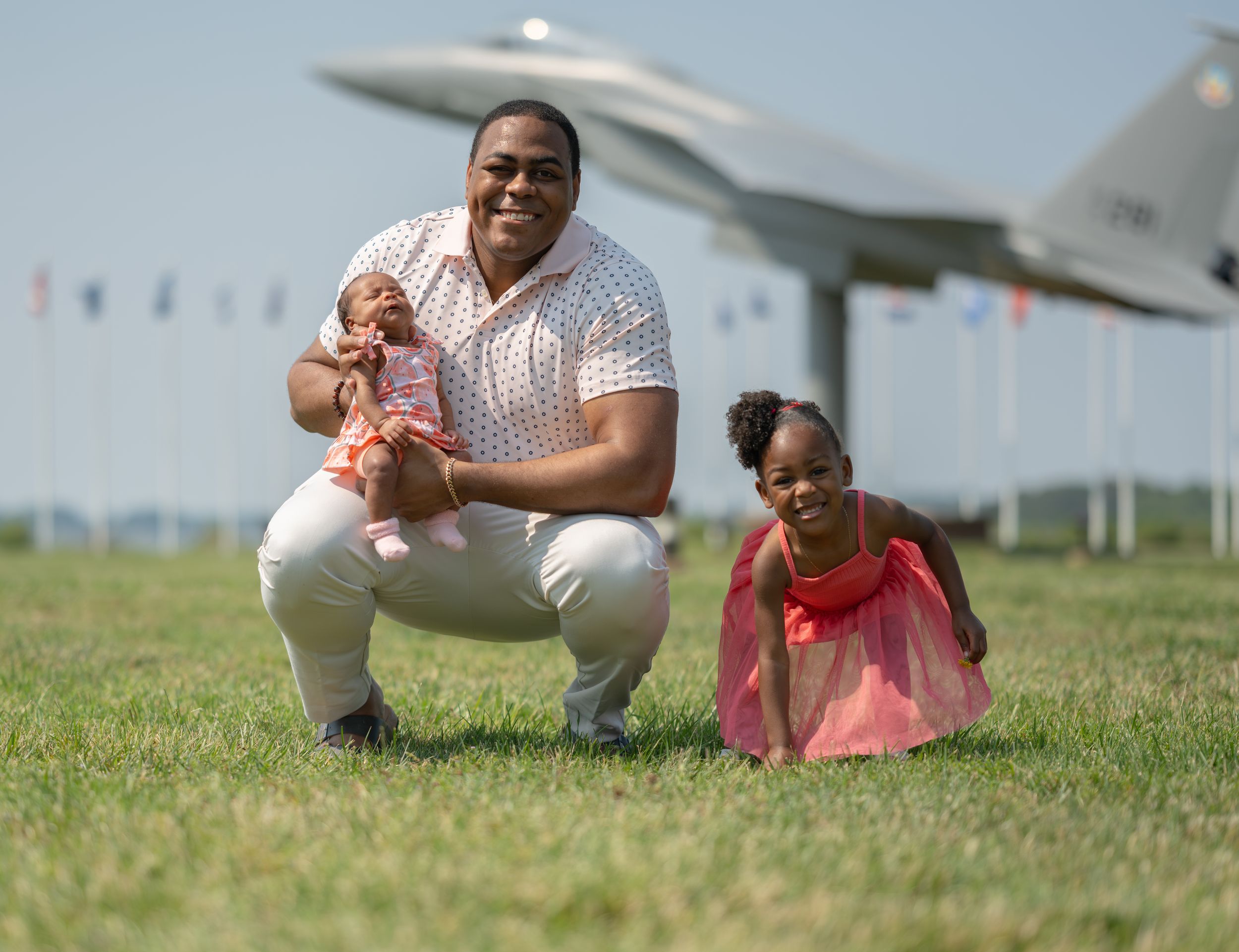 Two people posing together on grass near an aircraft display and flagpoles on a bright day.