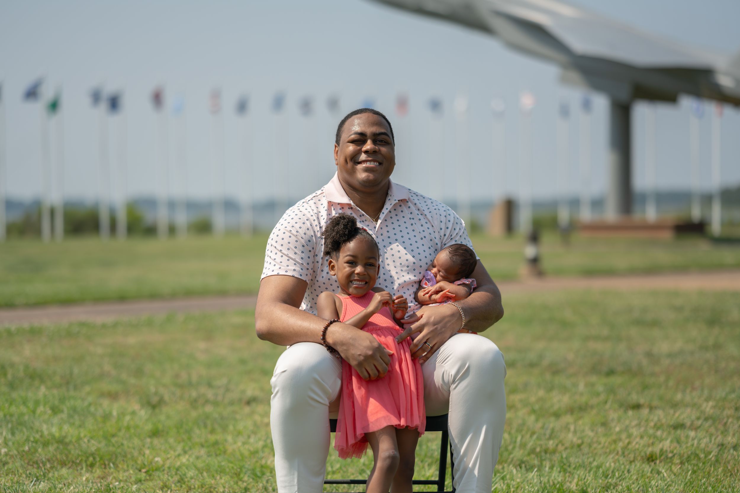 Person in pink shirt and white pants sitting cross-legged on grass near flagpoles.