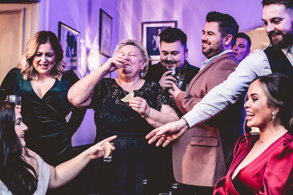 a grandmother takes a tequila shot with everyone watching her at a wedding