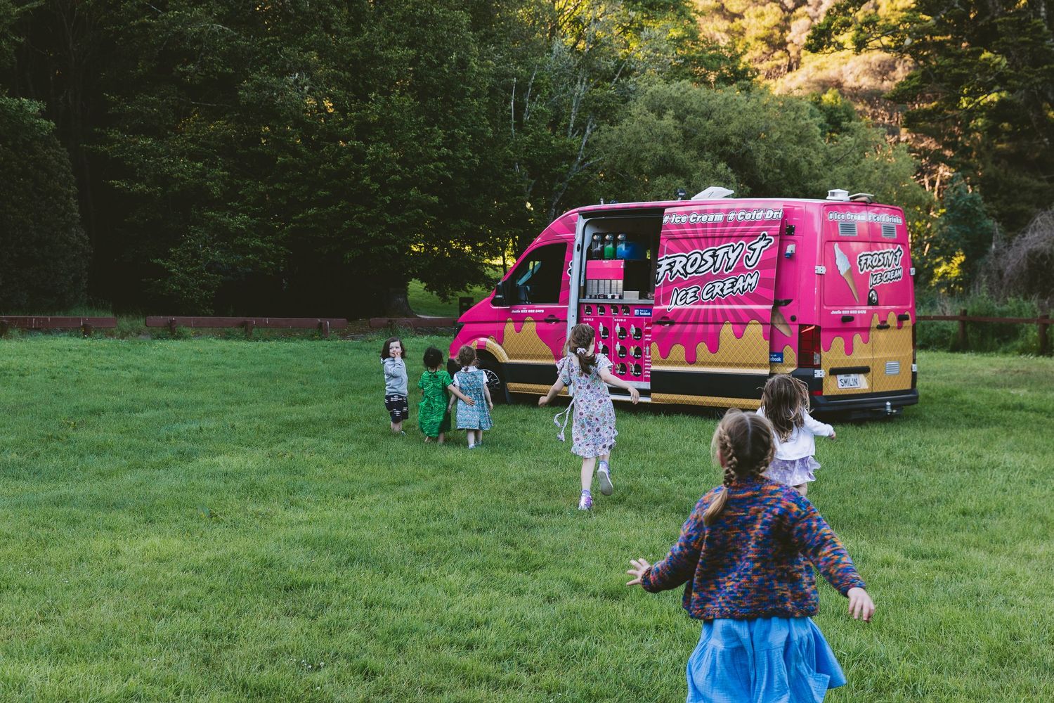 Children run toward a bright pink food truck parked on a grassy field at sunset.