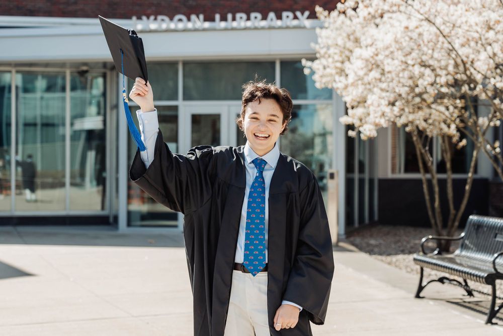 Graduate in academic regalia celebrates near library with cherry blossoms blooming.