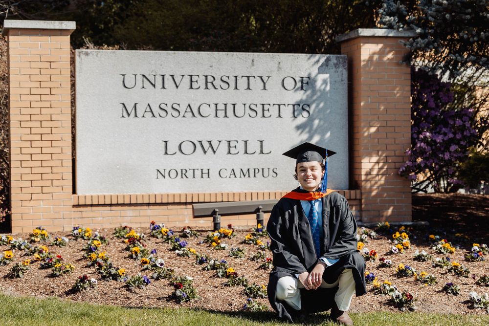 Graduate poses in front of University of Massachusetts Lowell North Campus sign during graduation.
