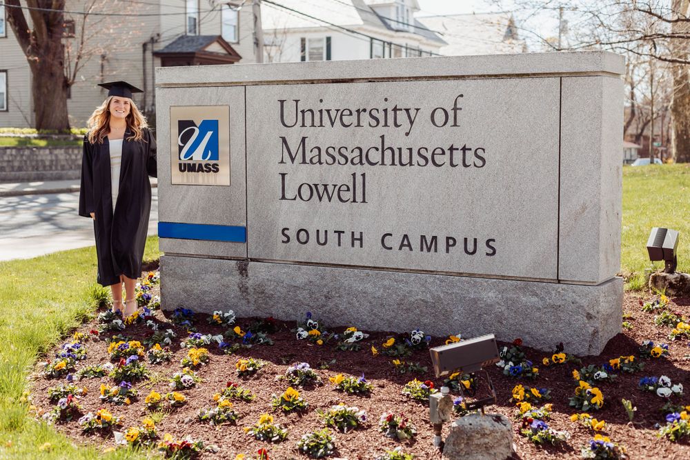 Graduate in cap and gown stands by UMass Lowell South Campus entrance sign with flowers.