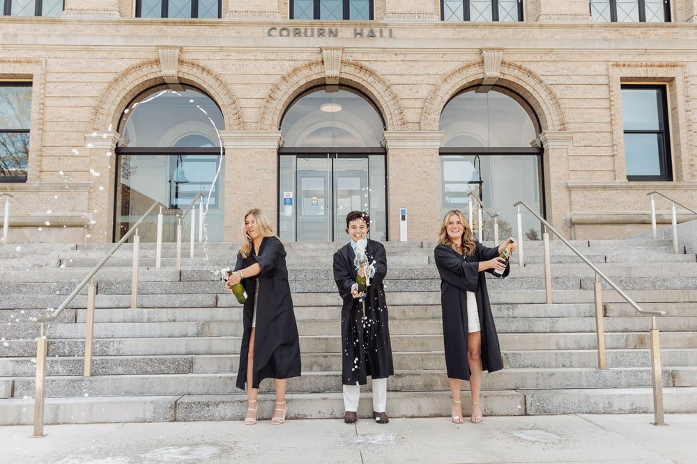 Three graduates in black graduation gowns pose on grand stone steps of university building.