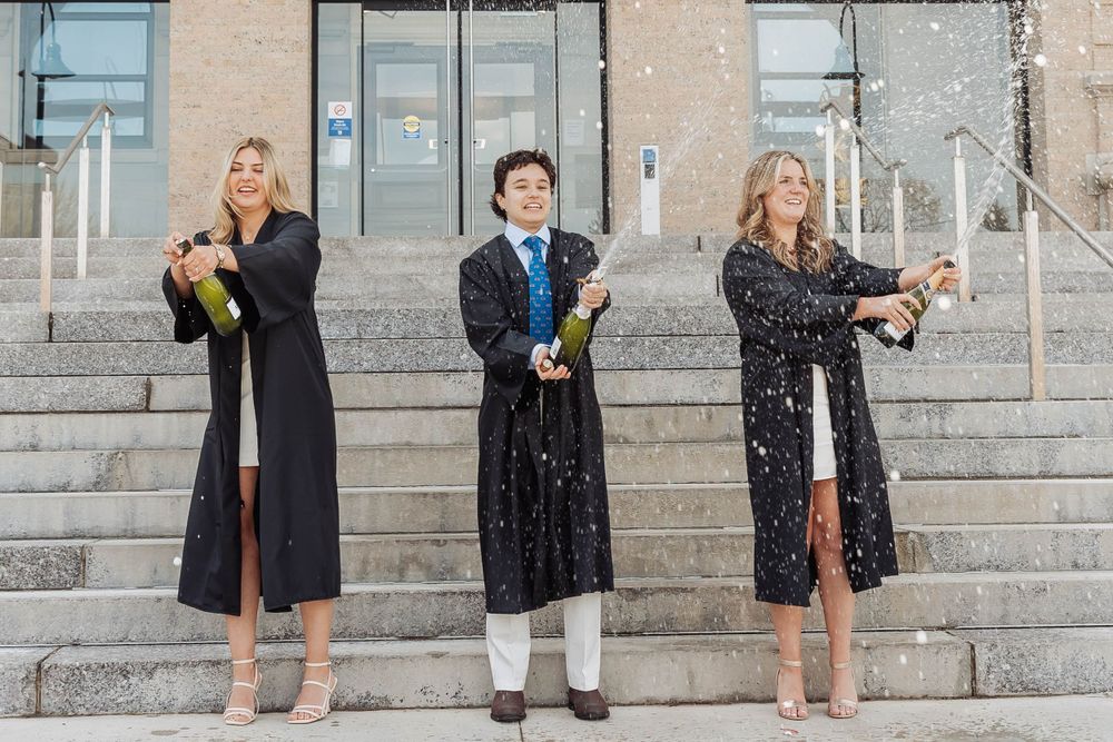 Three graduates in academic regalia pose with champagne bottles on university steps.