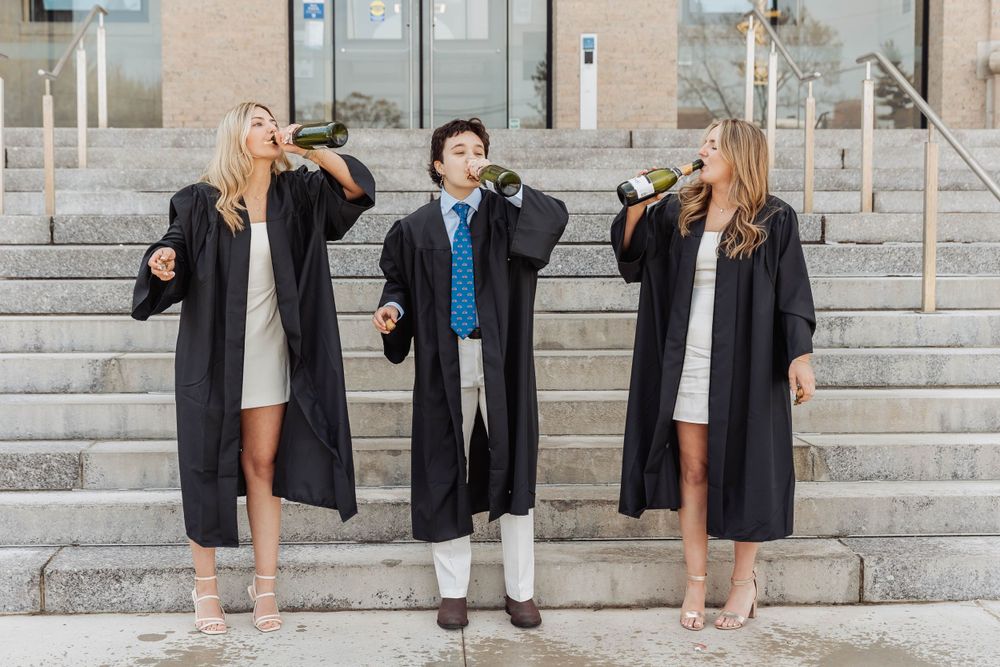 Three graduates in graduation attire playfully pose with champagne on university steps.
