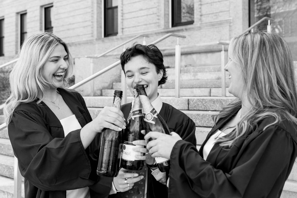 Three graduates toast with champagne bottles outside a stone building.