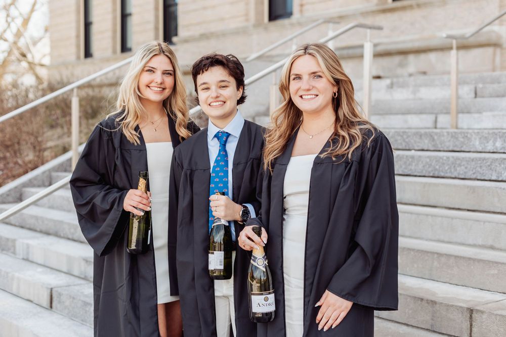 Graduates in black robes pose with champagne bottles on university steps.