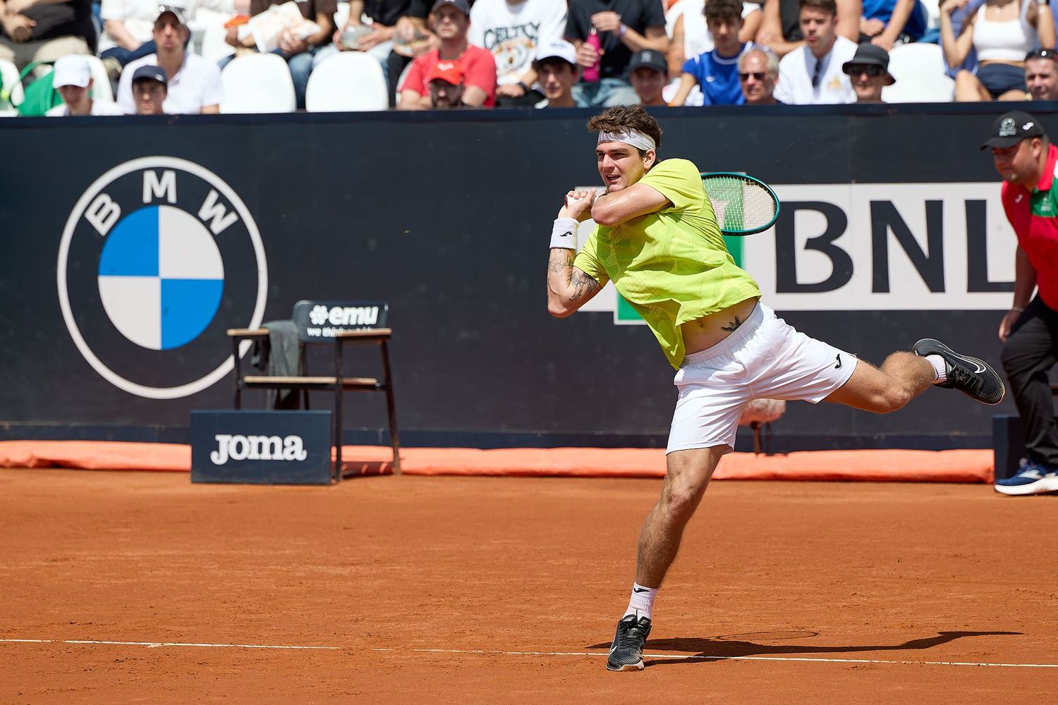 Tennis player in neon yellow shirt executing powerful serve during match on clay court.