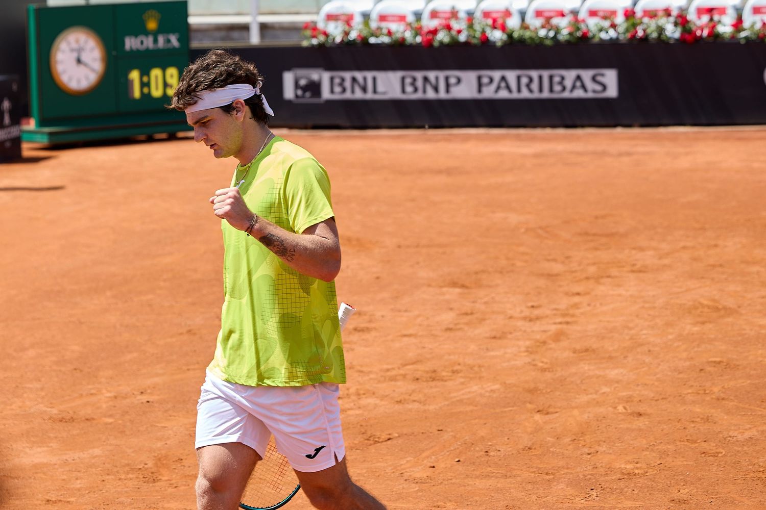 Tennis player in bright yellow shirt walking on clay court during tournament match.