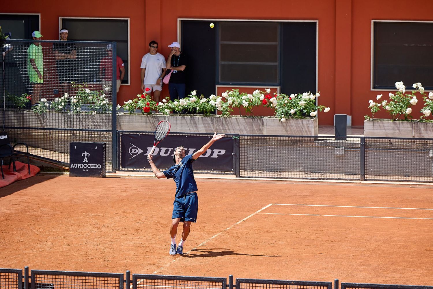 Tennis player serving during match on clay court with flower boxes and Dunlop advertising in background.