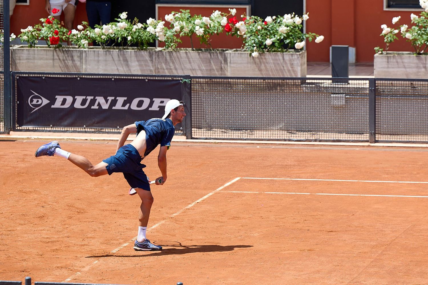 Tennis player executing powerful serve on clay court with Dunlop banner visible.
