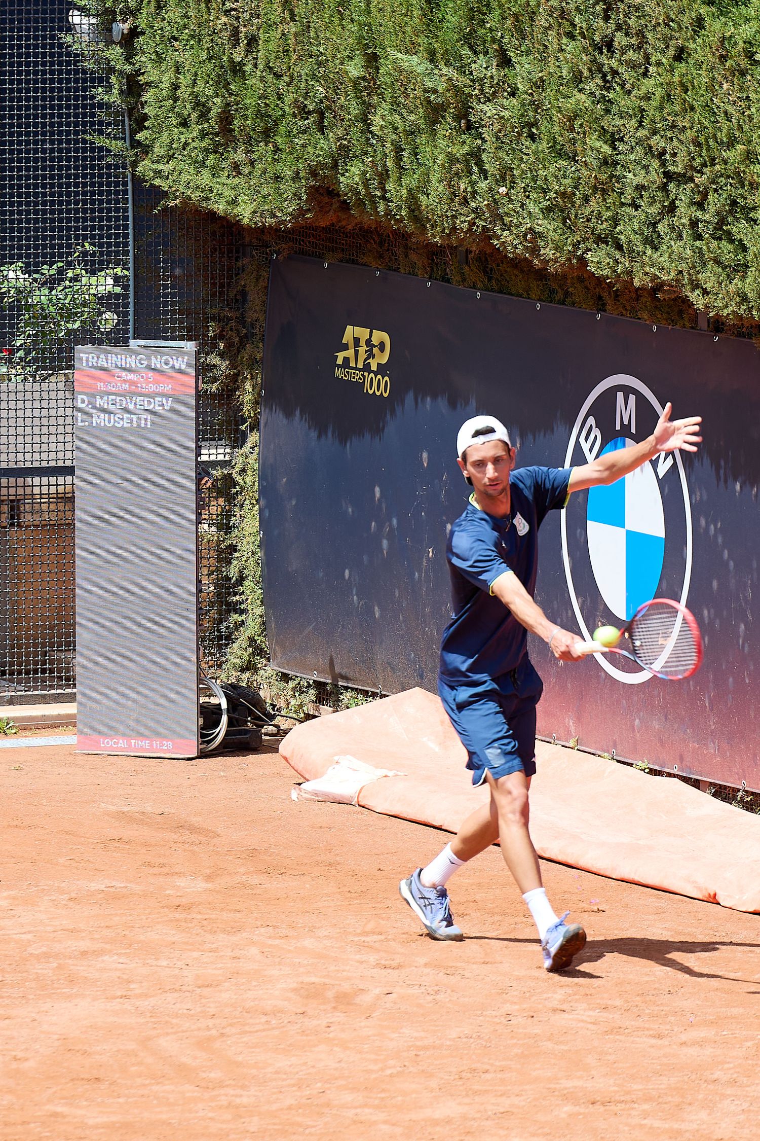 Tennis player hitting forehand shot on clay court with BMW logo visible in background.