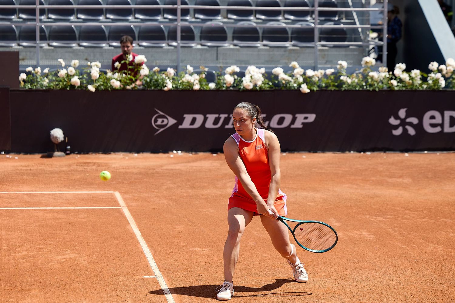 Tennis player preparing to return serve on clay court with white flowers decorating background.
