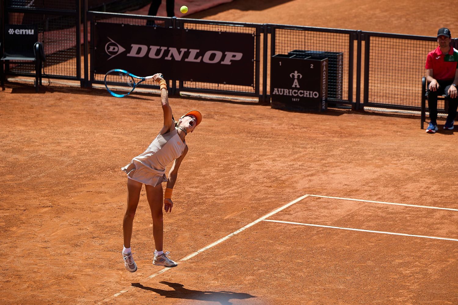 Tennis player serves on clay court with Dunlop banner visible in background during tournament match.