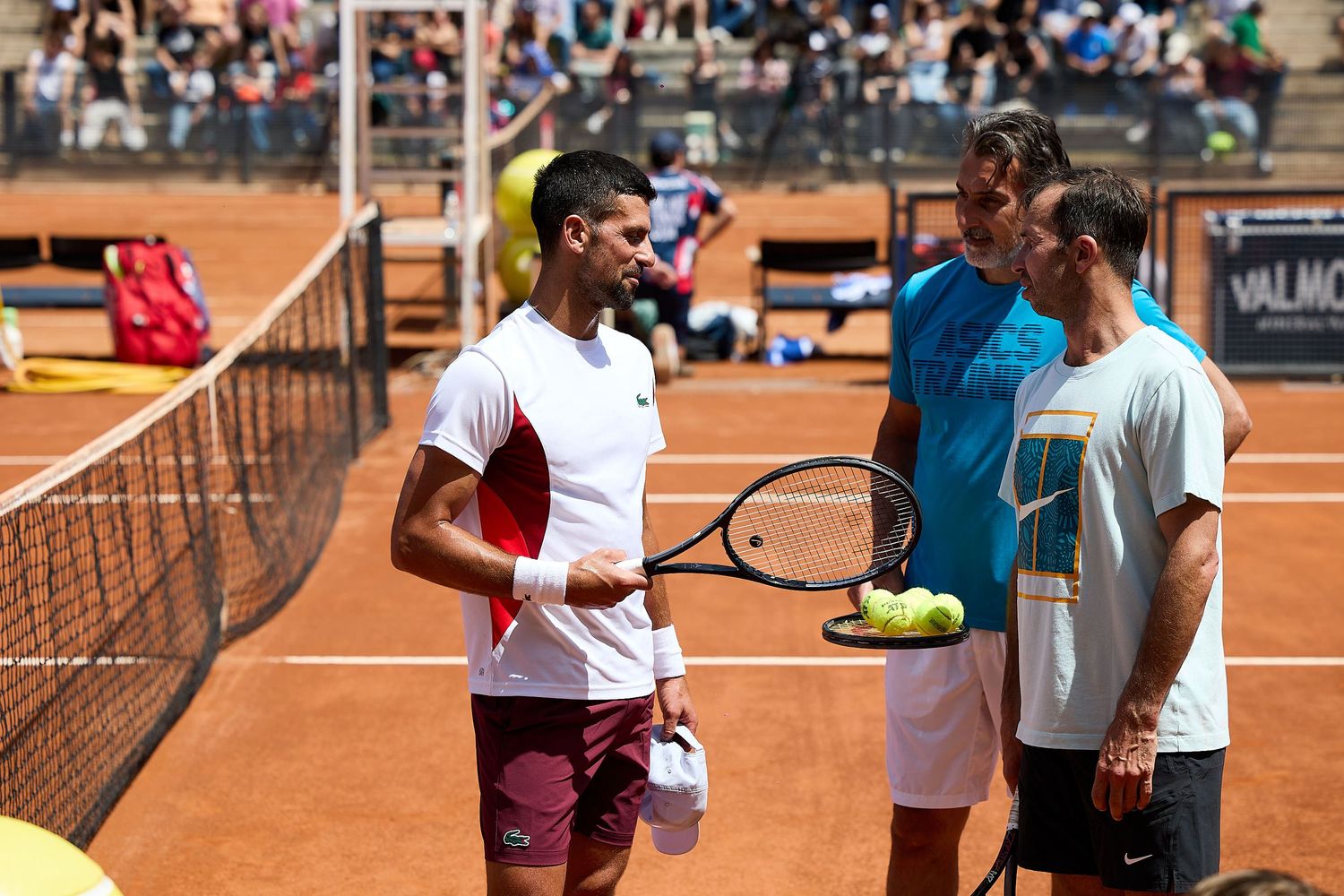 Tennis players meet at net on clay court after match with spectators in background.
