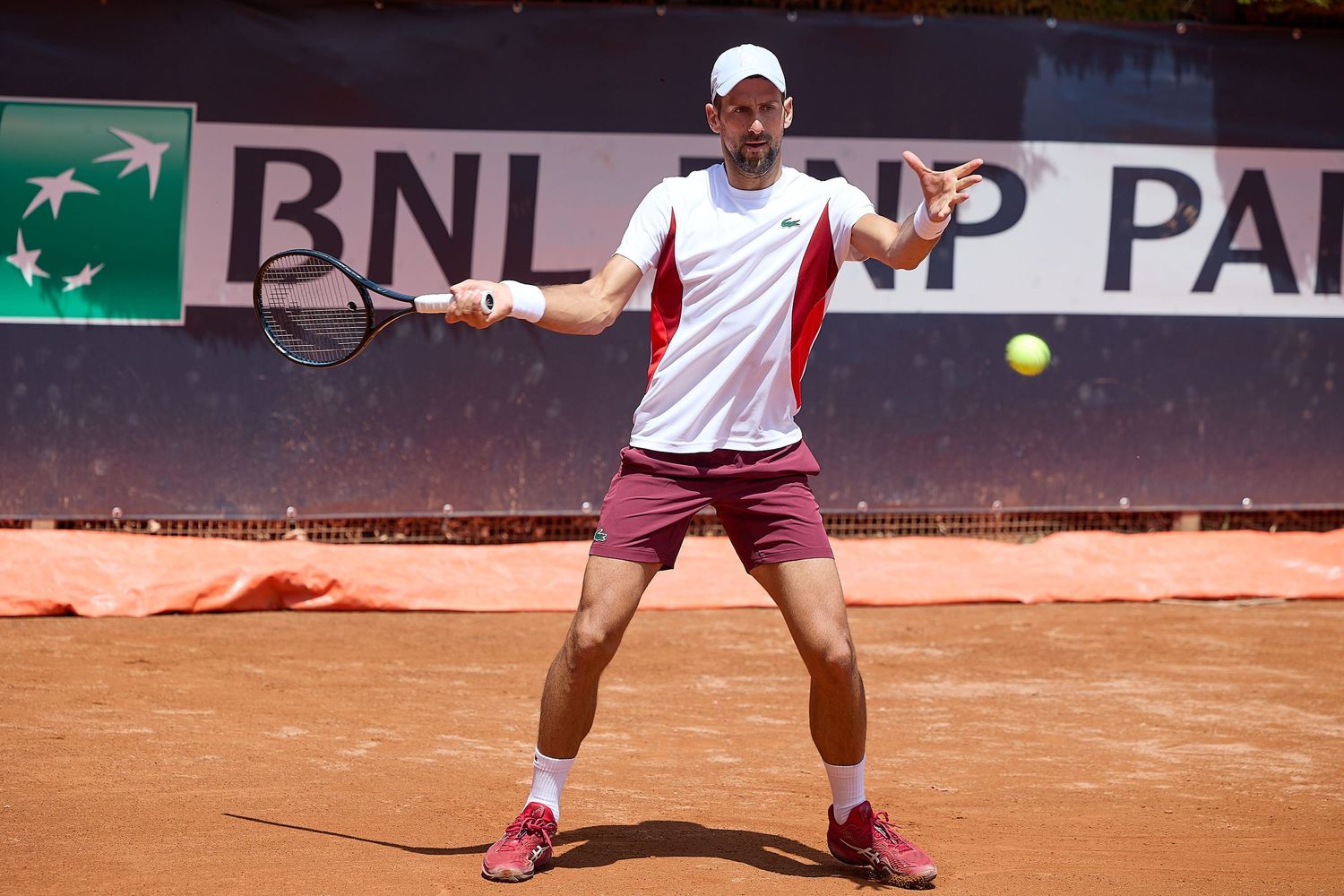 Tennis player in white and burgundy outfit returns ball on clay court during match.
