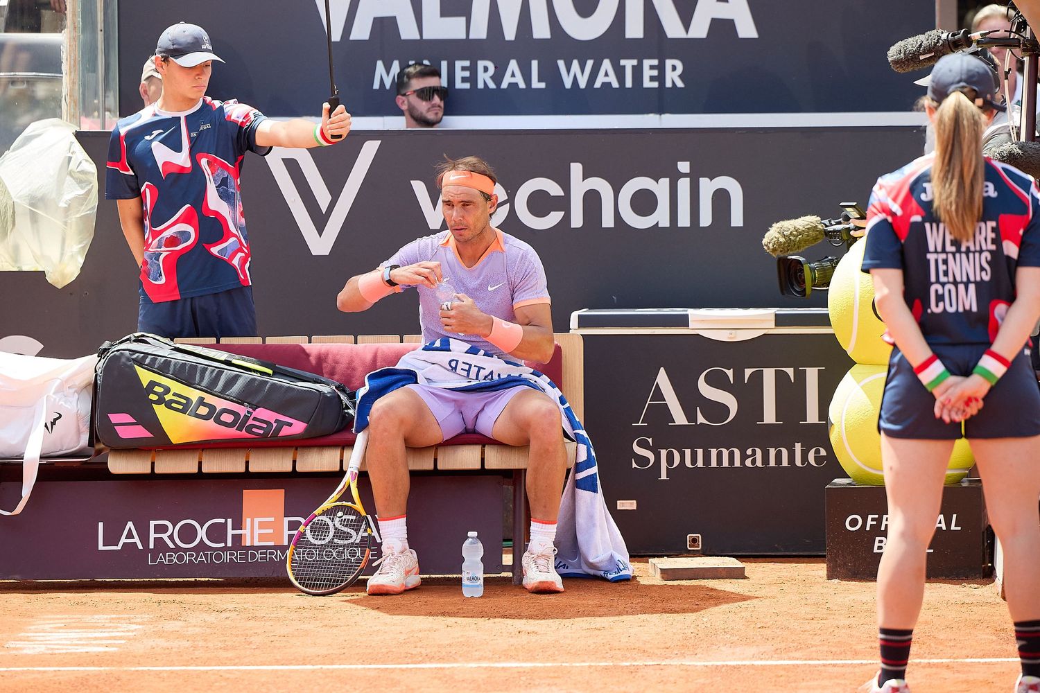 Professional tennis player rests on bench during clay court match at tournament.