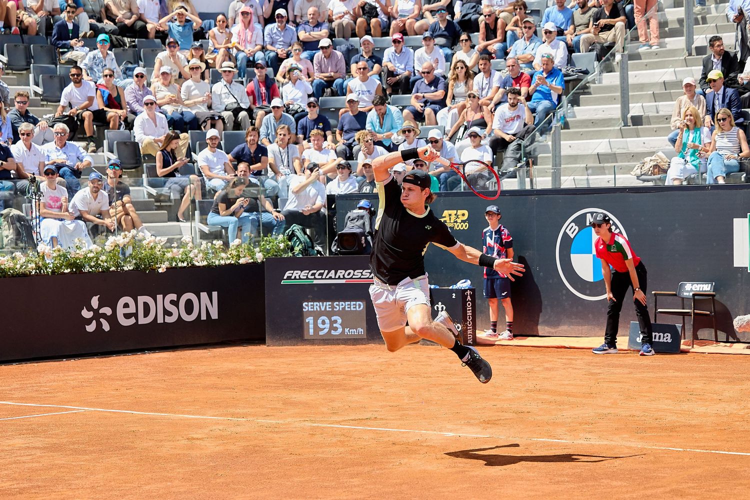 Tennis player leaps to return shot during clay court tournament match.