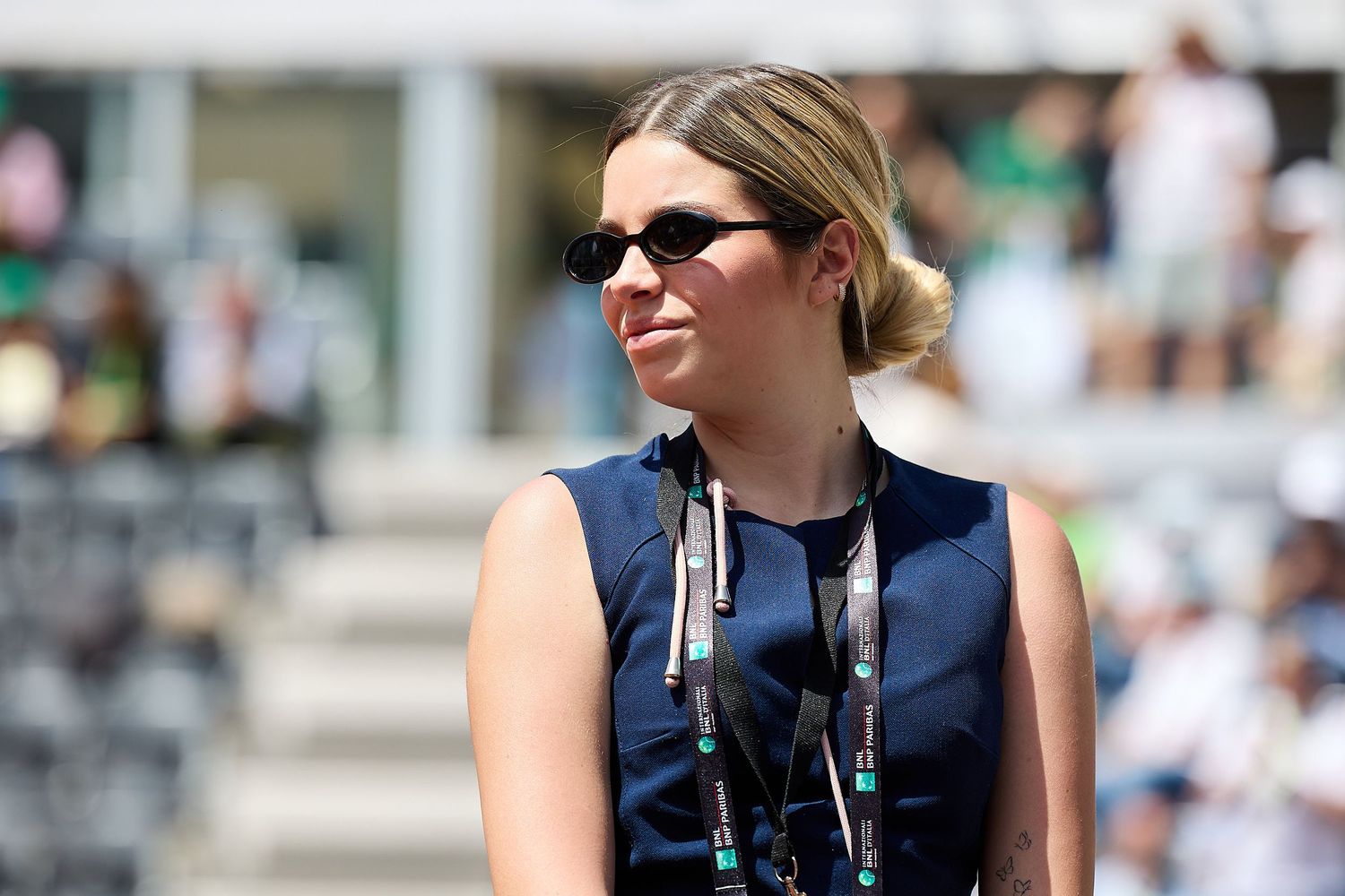 Event staff member wearing sunglasses and navy blue vest observes tennis match.