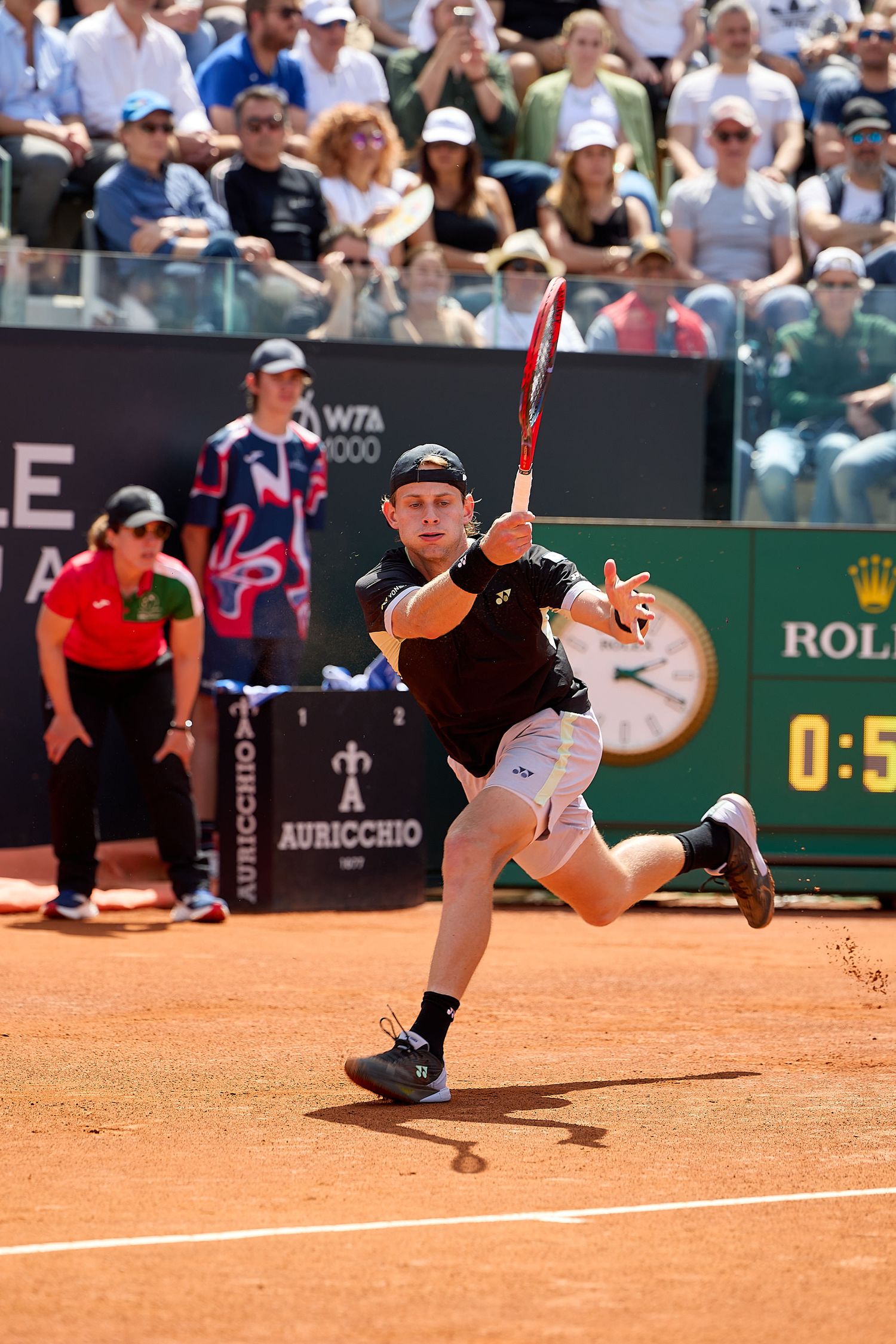 Tennis player in black shirt stretches for aggressive return shot on clay court.