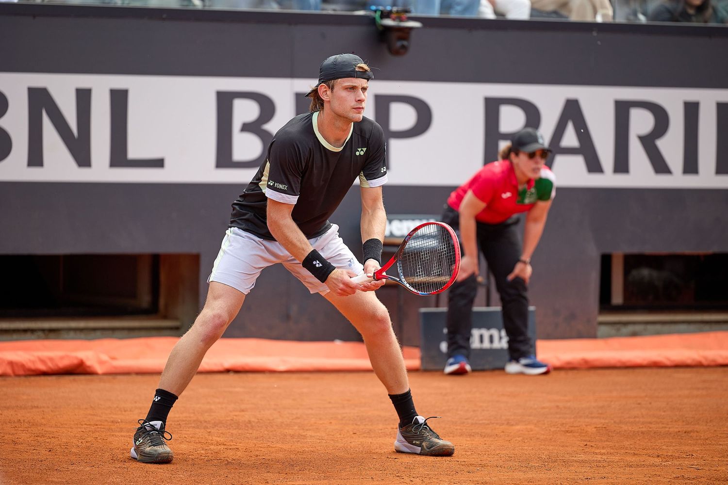 Tennis player in black shirt prepares to return serve during clay court match.