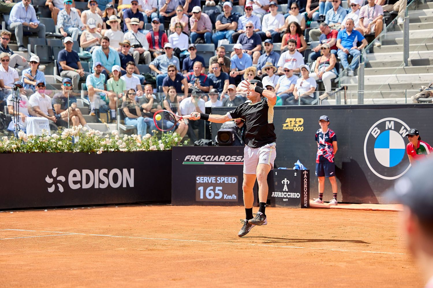 Tennis player serving on clay court with spectators and BMW branding visible.