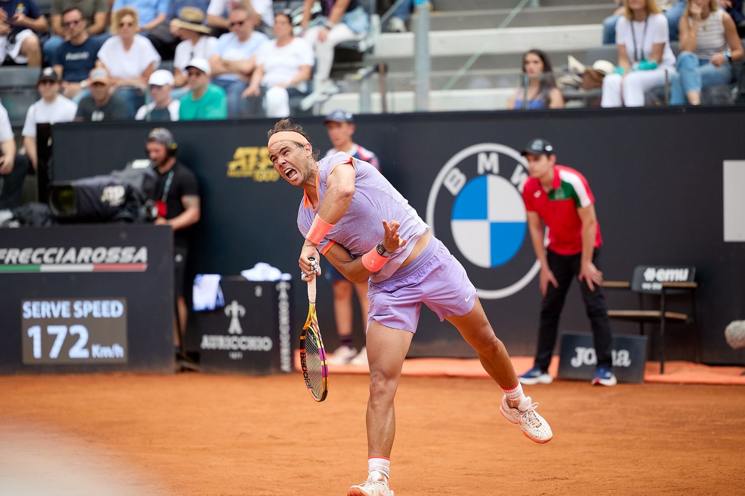 Tennis player serving on clay court with BMW logo visible in background.