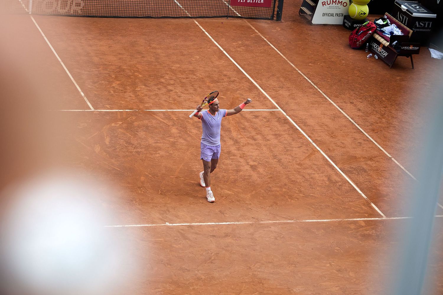 Overhead view of tennis player serving on clay court during match.