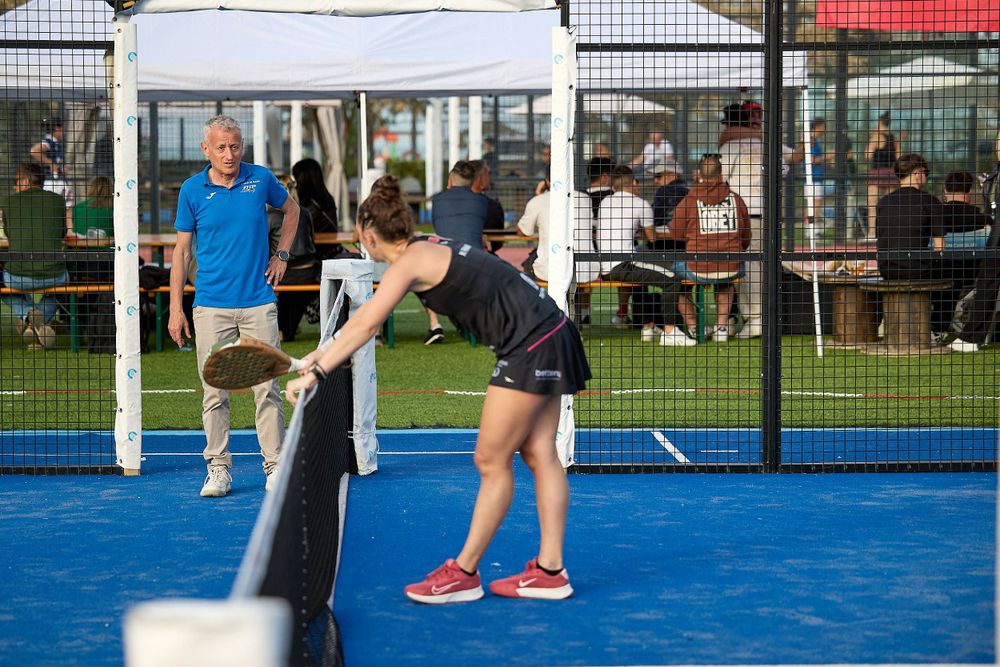 Player bending down on blue padel court surface during game match with spectators in background.