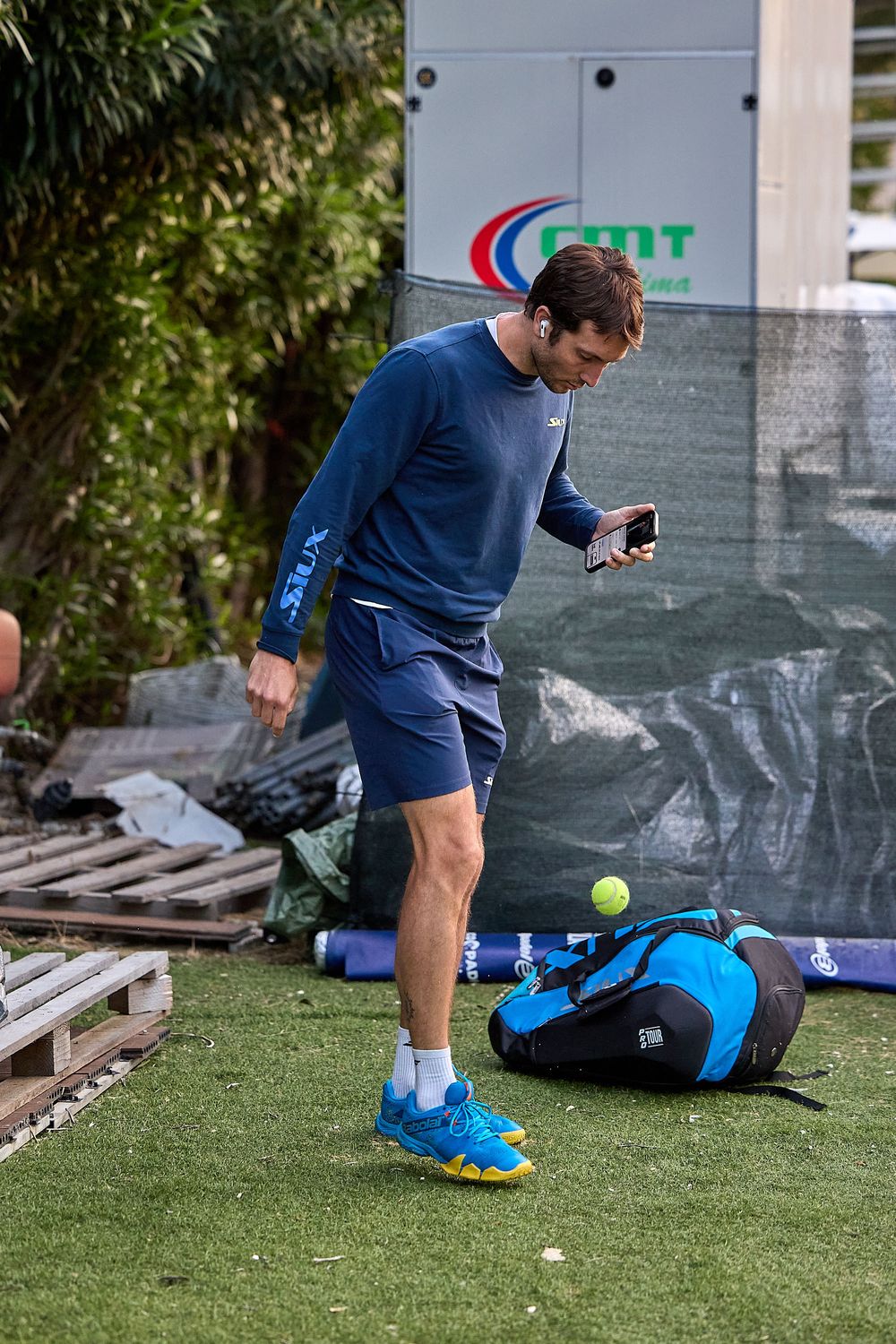 Athlete checking phone on artificial turf next to tennis bag and equipment.