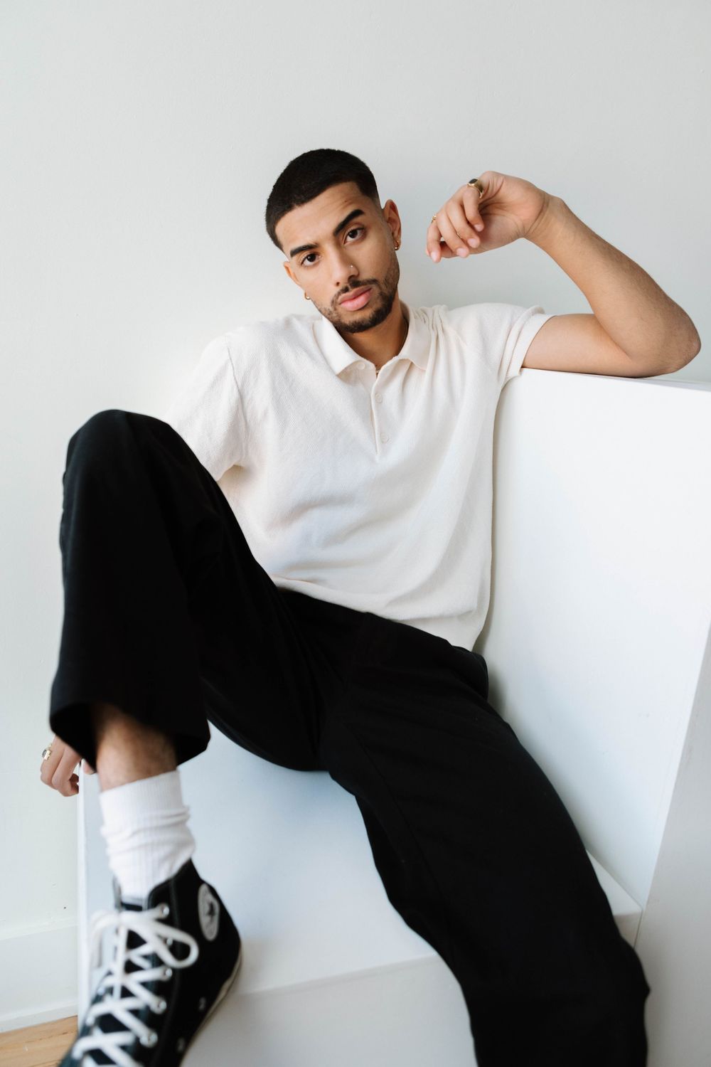 A person in a white polo shirt and black pants lounges casually against a white wall while wearing black sneakers.