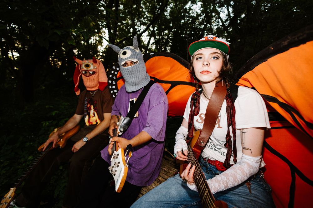 Three musicians with halloween masks perform outside by orange tents during a camping trip.