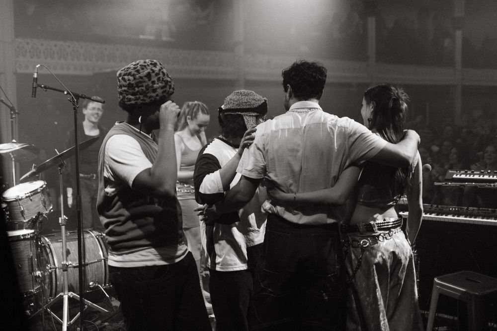 Black and white photo showing musicians on stage huddled together during a performance with instruments and microphones.