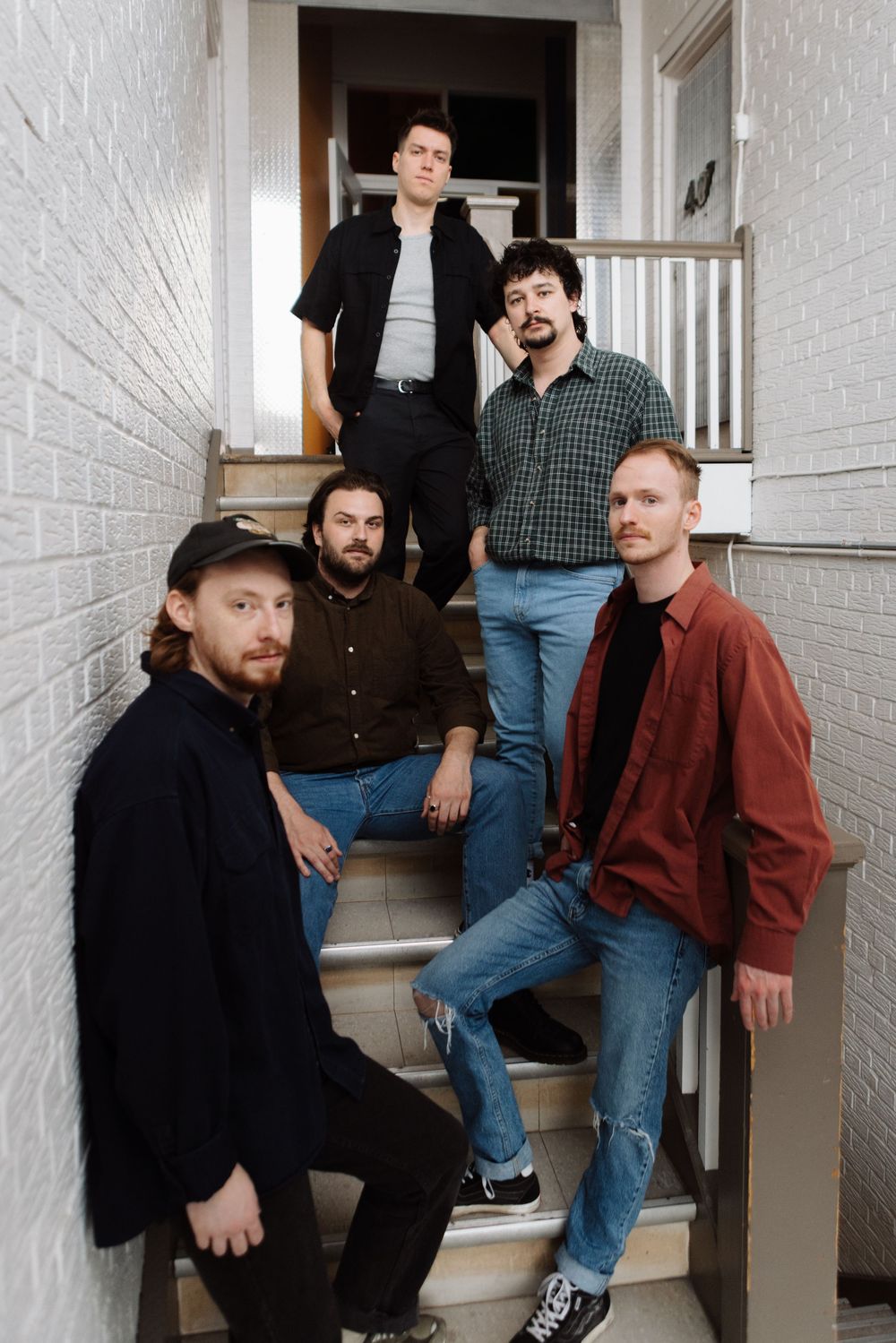 Five musicians pose on a white staircase wearing casual clothing in an intimate indoor setting.