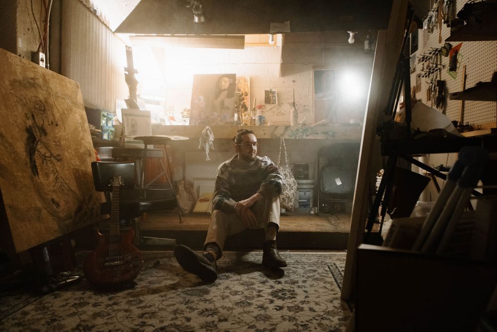 A figure sits in a dimly lit art studio or workshop space, surrounded by tools and equipment with sunlight streaming in.
