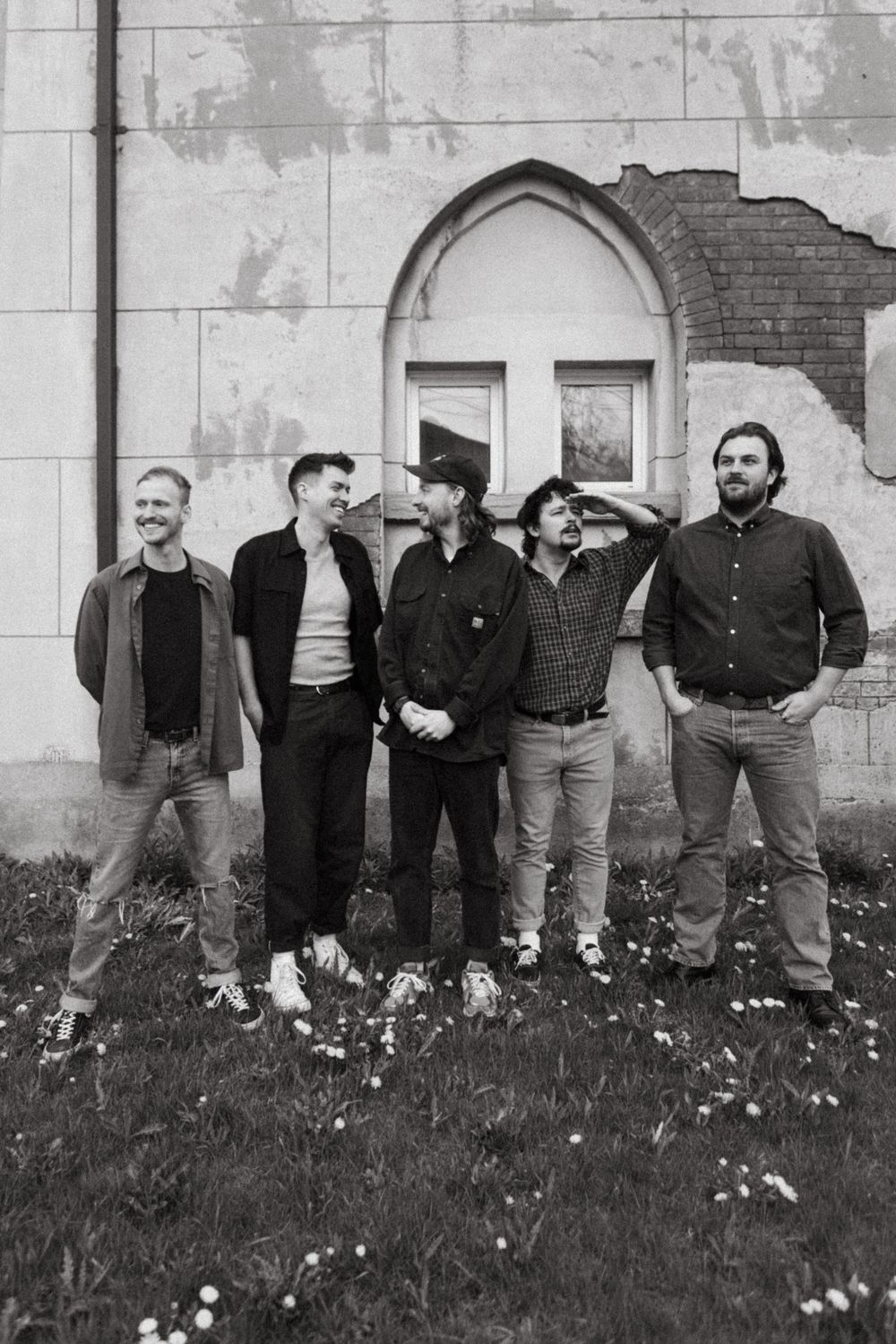Black and white photo of five band members standing together against a stone wall with an arched architectural detail.