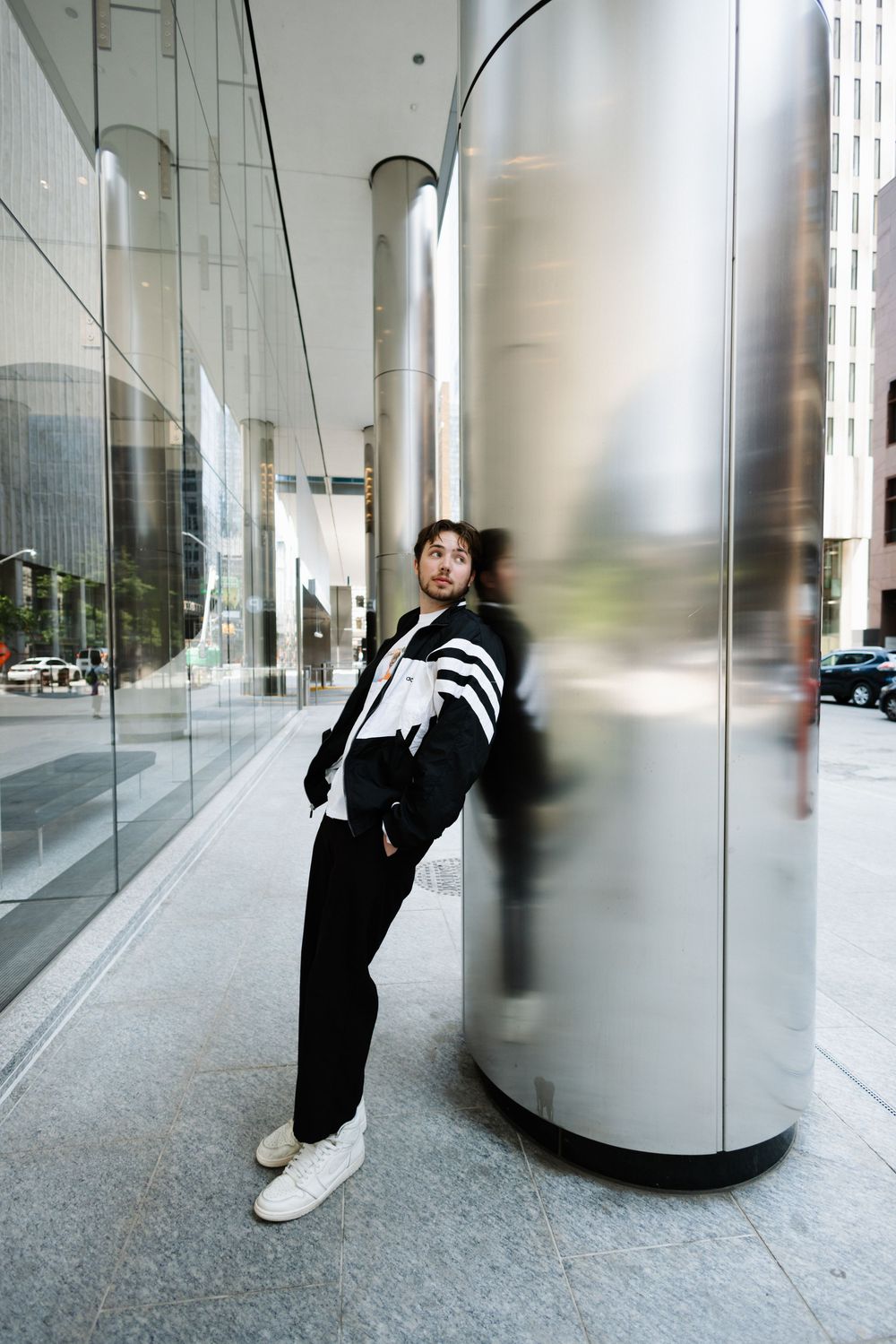 A person in a black and white athletic jacket leans against a metallic column on a modern city street.