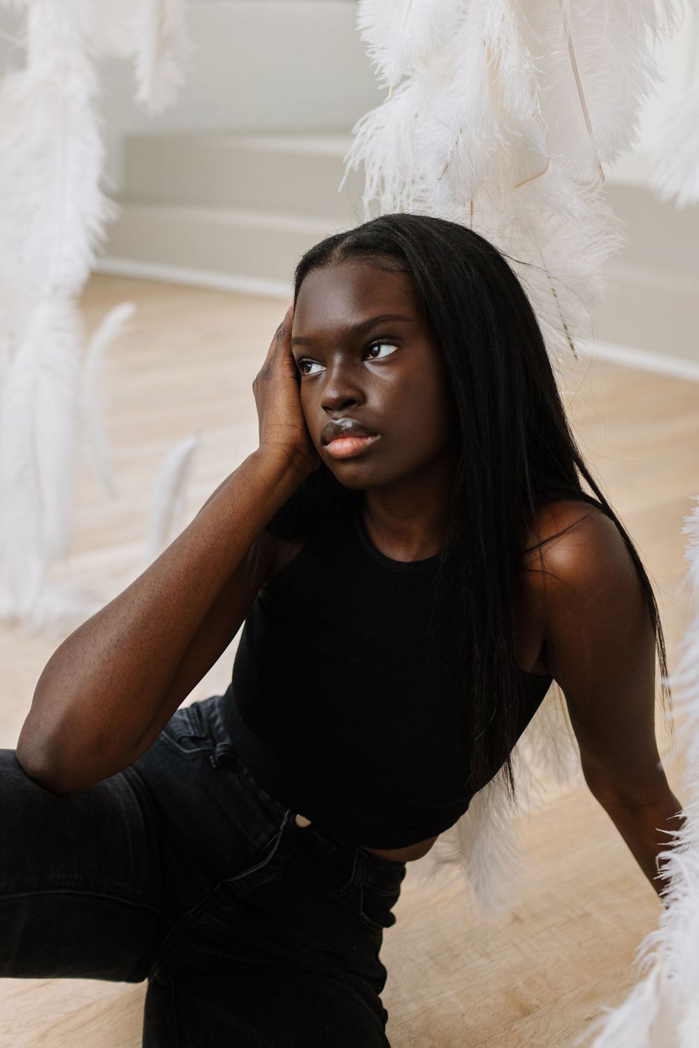 Artistic portrait surrounded by white feathers captures contemplative pose against light wooden floor.