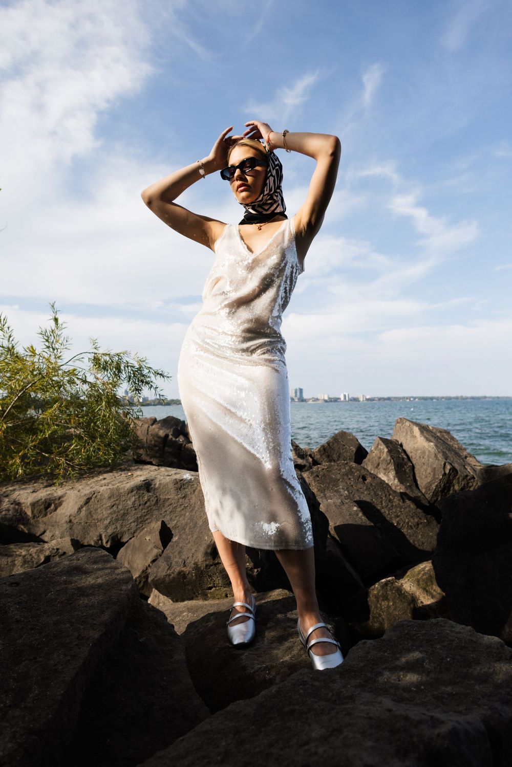 Figure in flowing white dress and sunglasses poses on rocky shoreline with blue water and skyline visible in background.