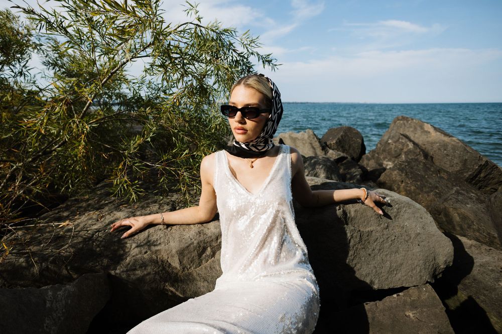 Person in white dress and sunglasses sits on rocky beach shore with ocean and blue sky in background.