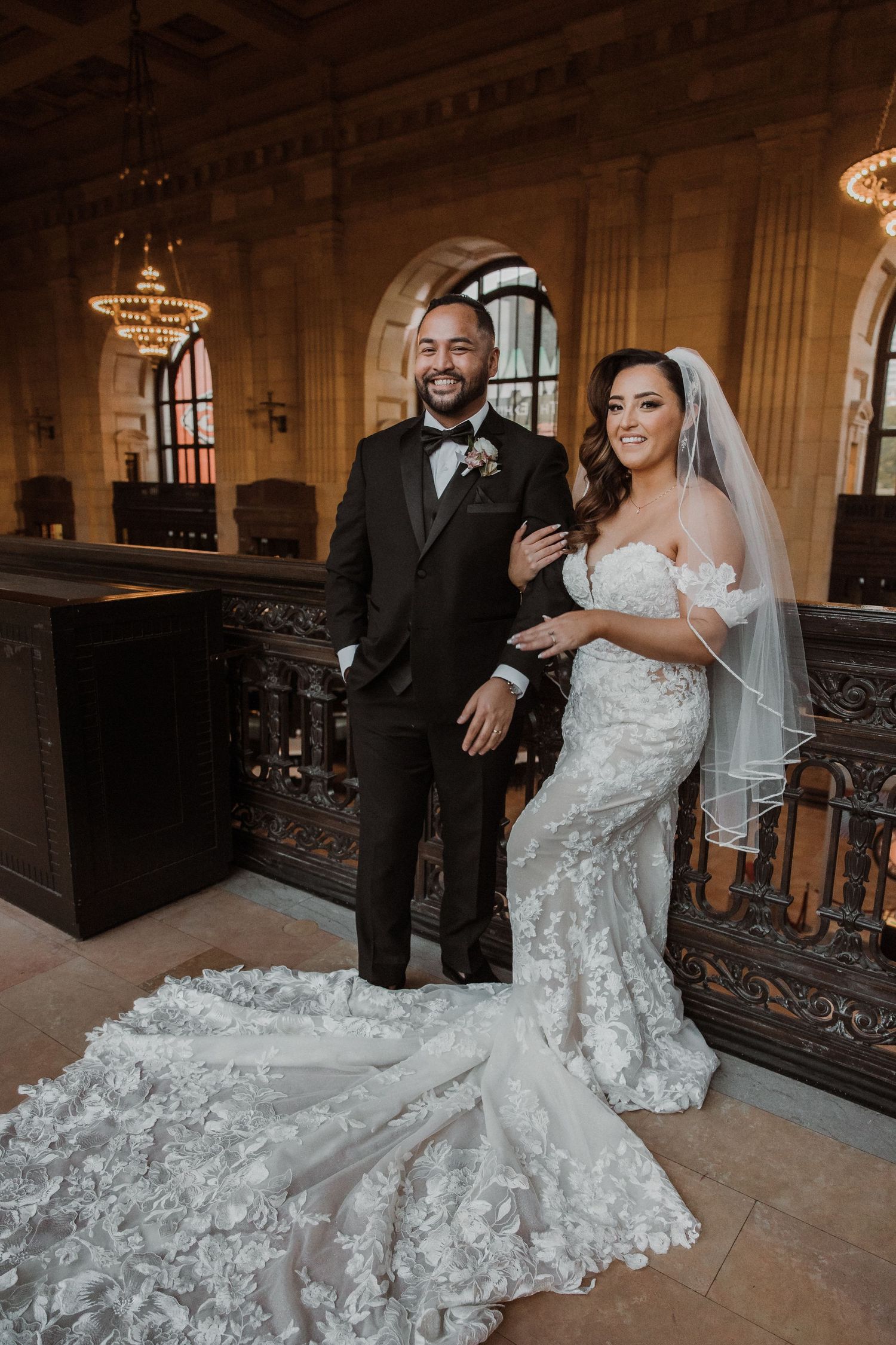 Elegant wedding couple posing in grand ballroom with ornate chandeliers and architecture.