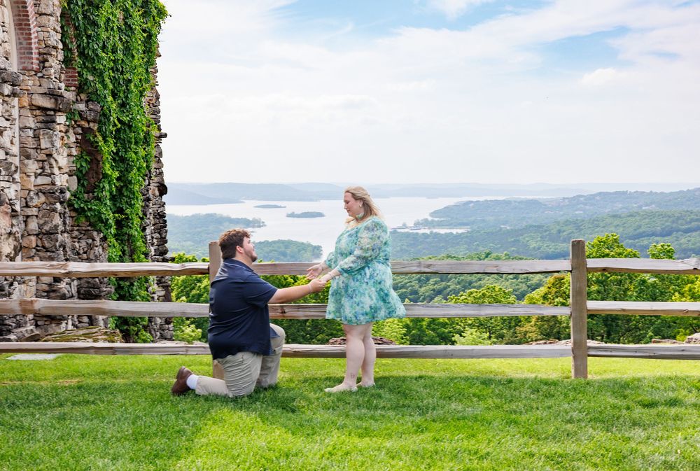 A romantic proposal moment takes place on a scenic mountain overlook with green grass and a rustic stone wall.