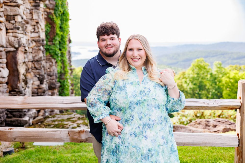 A couple poses together outdoors on a mountain overlook with stone ruins and greenery in the background.