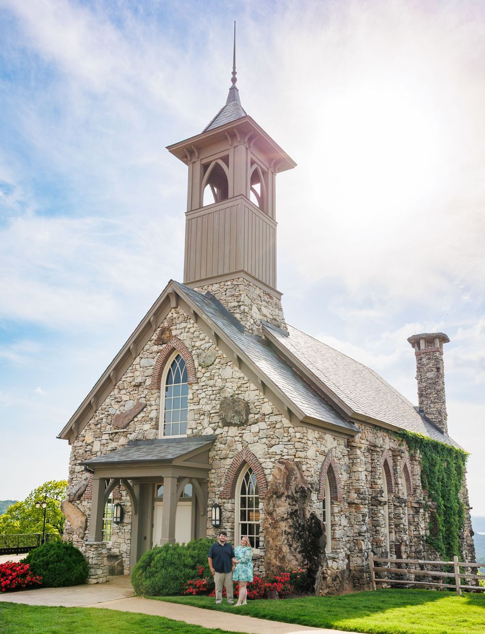 Historic stone church with white bell tower and arched windows stands against a blue sky in a scenic countryside setting.