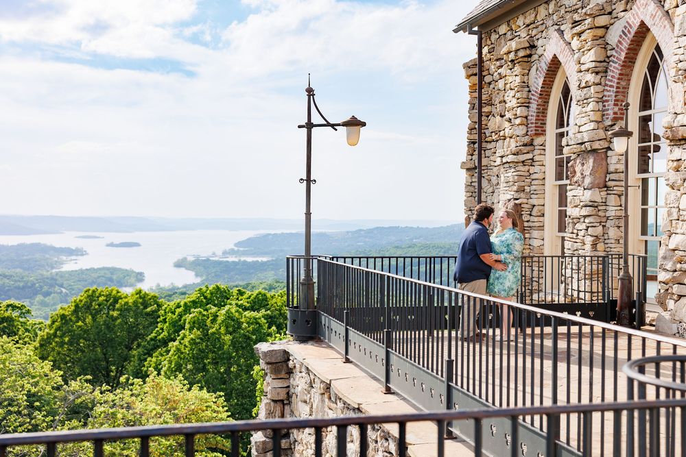 Two people enjoy a scenic mountain view from a stone building's balcony overlooking lakes and rolling hills on a sunny day.