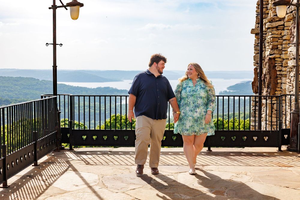A couple walks together at a scenic mountain overlook with stone architecture and railing during a sunny day.