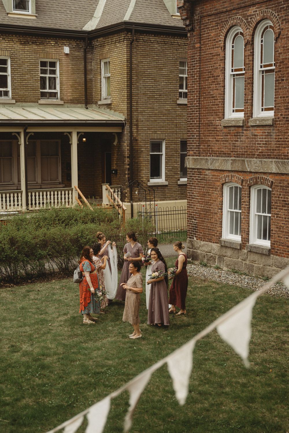 A small group gathers on the lawn outside a brick Victorian-style building with white bunting decorations.