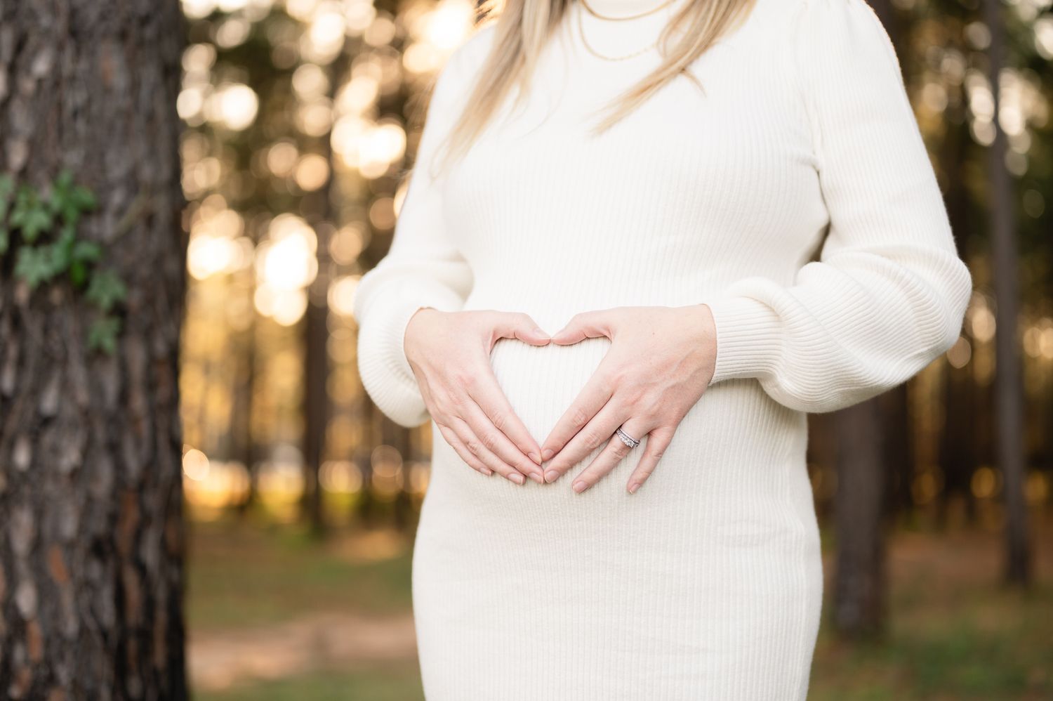 A close-up of hands forming a heart shape over a white sweater-clad baby bump in a forest setting.