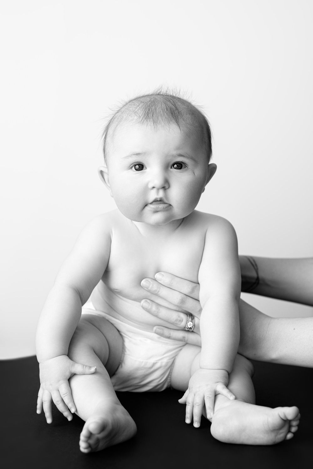 Black and white studio portrait of baby in diaper sitting against white background.